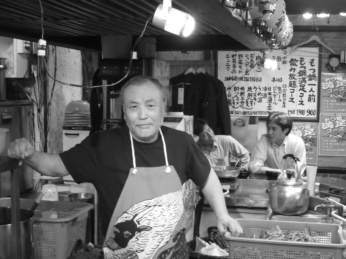日本の居酒屋の厨房で、エプロンをつけた男性がカメラを見ている白黒写真。背景には客らしき男性たちや、もつ鍋や飲み放題のメニューが書かれた掲示が見え、賑やかな飲食店の雰囲気が伝わる。