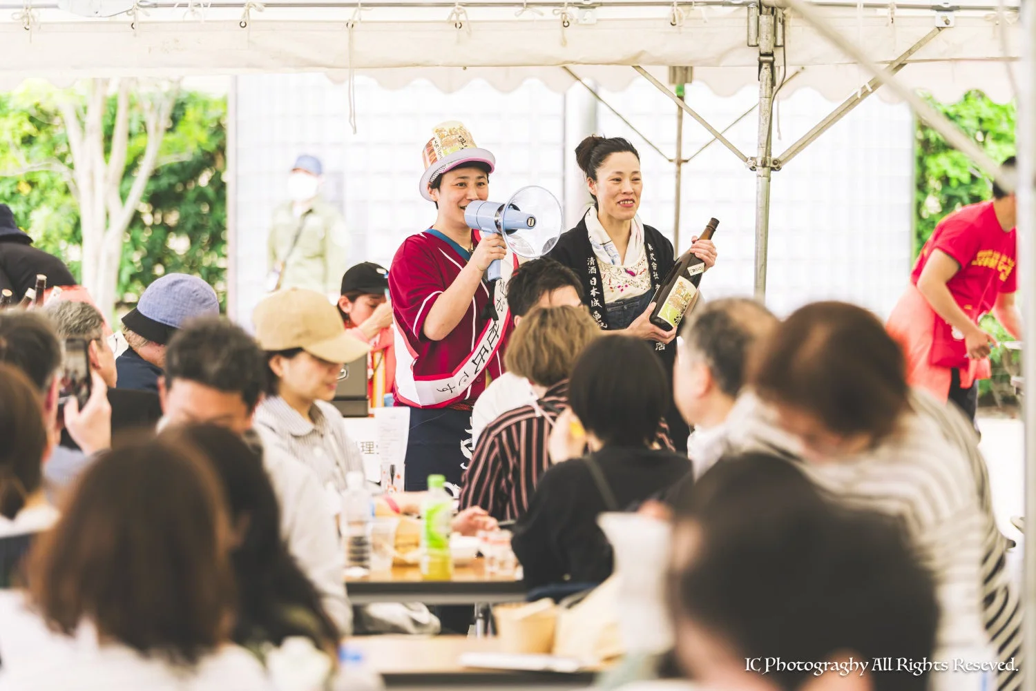 屋外のテントの下で、2人の女性が聴衆に向けて日本酒のプレゼンテーションを行っている様子