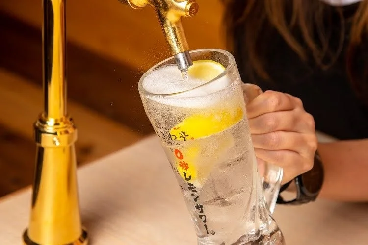 黄金色のサーバーから、氷とレモンスライスが入ったグラスに冷たいレモンサワーが注がれている瞬間