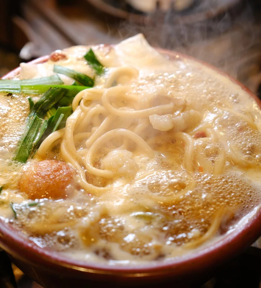 土鍋で煮立つ熱々の麺料理のクローズアップ。太麺と具材が白いスープの中で泡立ち、湯気が立ち上っている。食欲をそそる温かい一品。