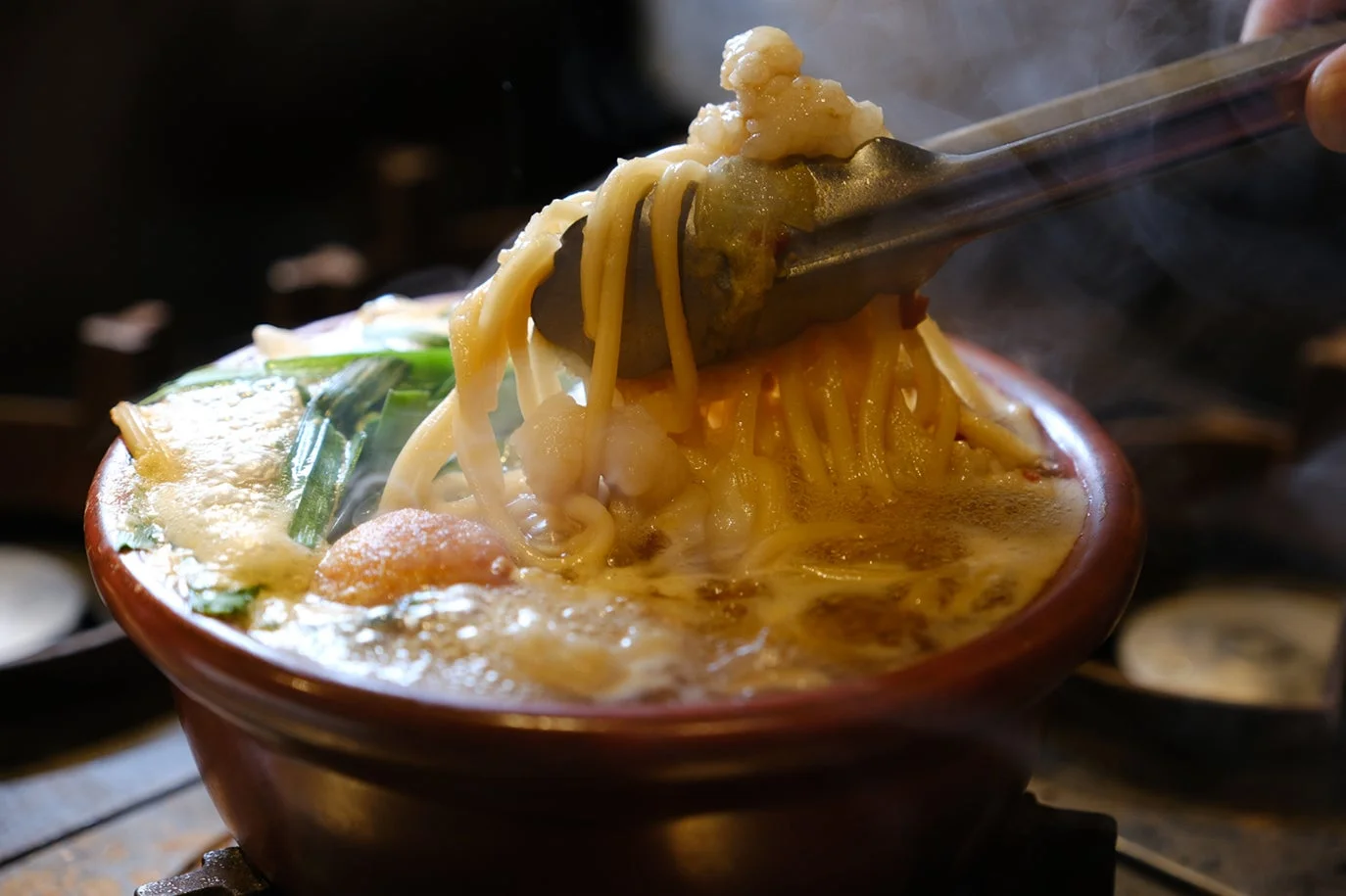 湯気を立てて煮込まれた鍋料理のクローズアップです。トングで持ち上げられた太めの麺と具材が確認でき、食欲をそそる温かい一品であることがうかがえます。