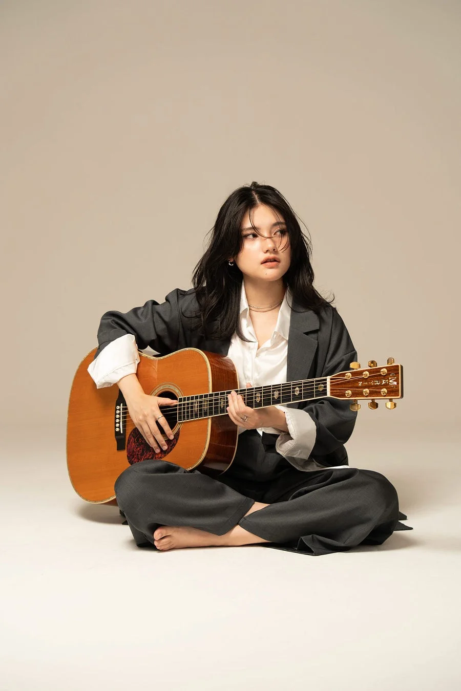グレーのジャケットと白いシャツを着た若い女性がアコースティックギターを演奏しているポートレート