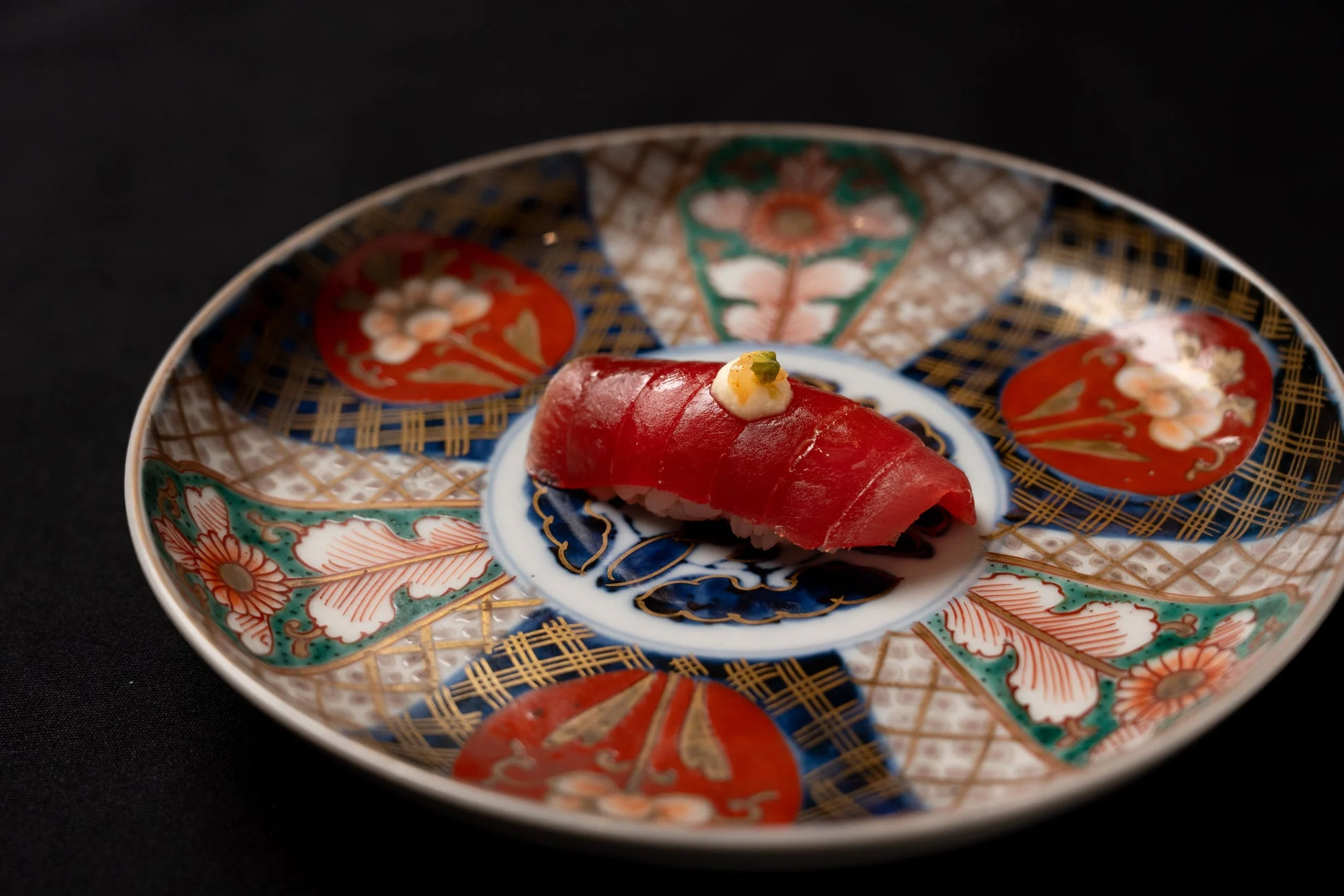 豪華な絵柄の皿に盛り付けられた、赤身のマグロの握り寿司。白いソースと緑の薬味が添えられ、洗練された一品です。