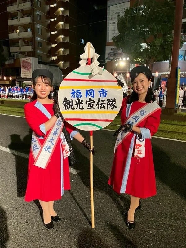 夜のイベントで「福岡市 観光宣伝隊」の看板を持つ大使