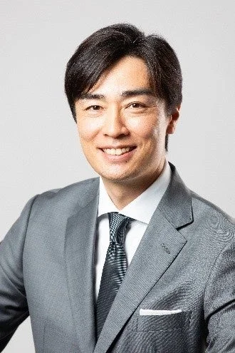 グレーのスーツを着たアジア人男性が、白いシャツと柄物のネクタイを着用し、カメラに向かって優しく微笑んでいます。真面目さと親しみやすさを兼ね備えた印象のビジネスポートレートです。