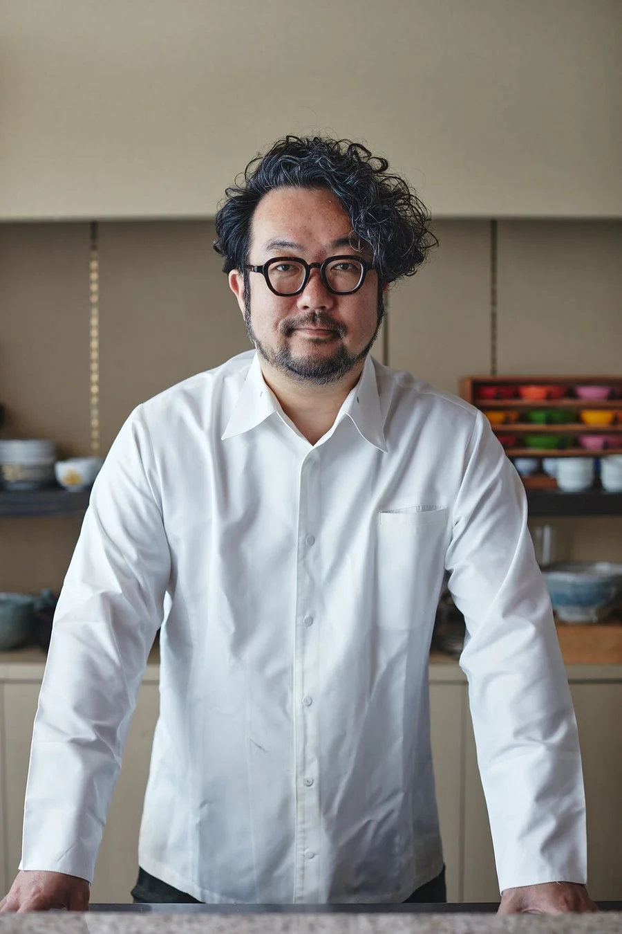 白いシャツを着てメガネをかけた男性が、カメラ目線で立っているポートレート写真です。背景にはキッチンを思わせる棚と食器が見えます。