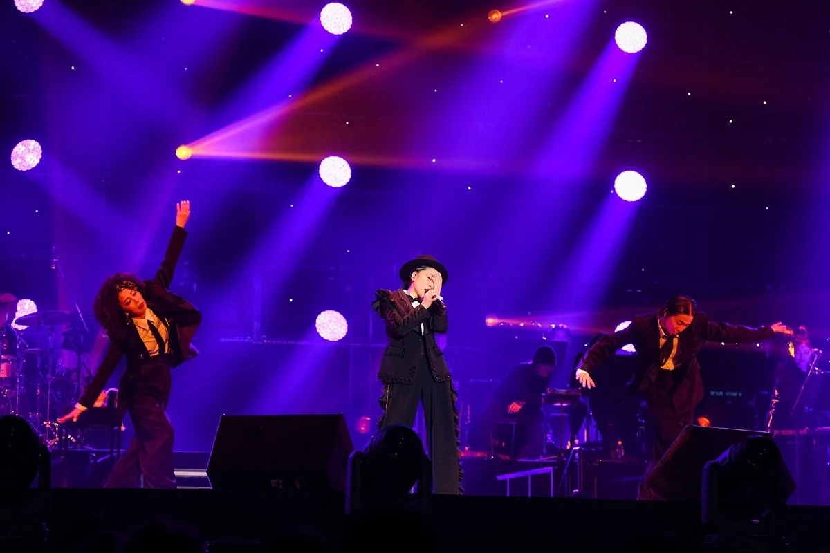 紫と青の照明が照らすステージで、中央の歌手と両脇のダンサーが情熱的なライブパフォーマンスを繰り広げている様子。