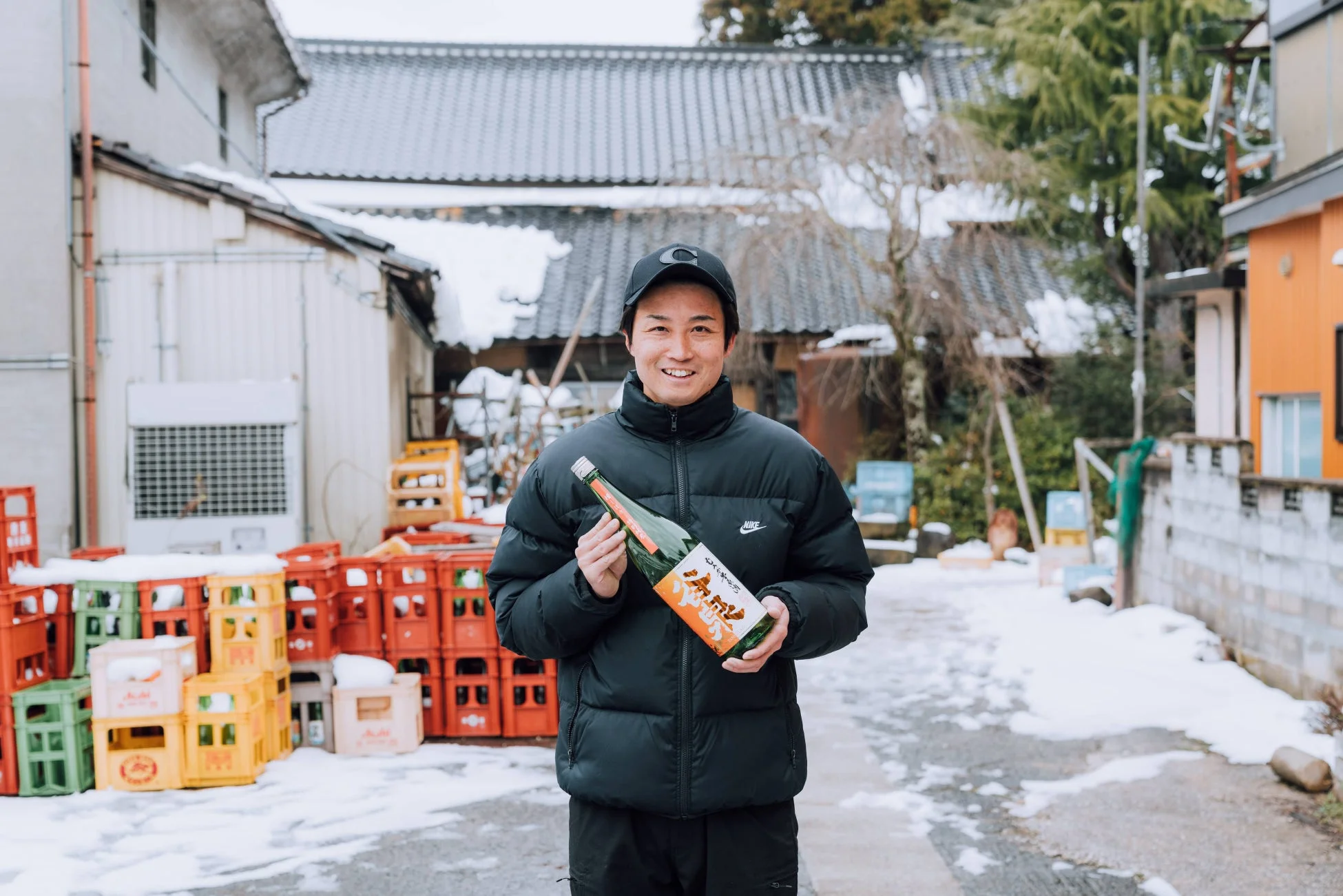 雪が積もる道で、黒いダウンジャケットを着た男性が笑顔で「天狗舞 山廃仕込 純米酒」のボトルを抱えている様子