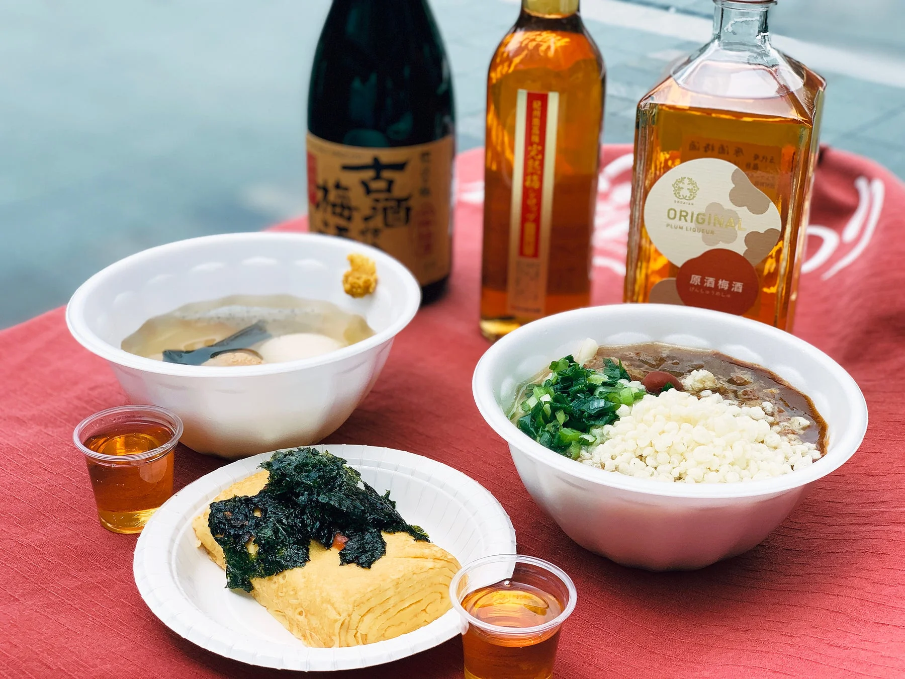 おでん、海苔がのった卵焼き、カレーうどんといった日本食が、複数の梅酒のボトルと共に赤い布の上に並べられています。