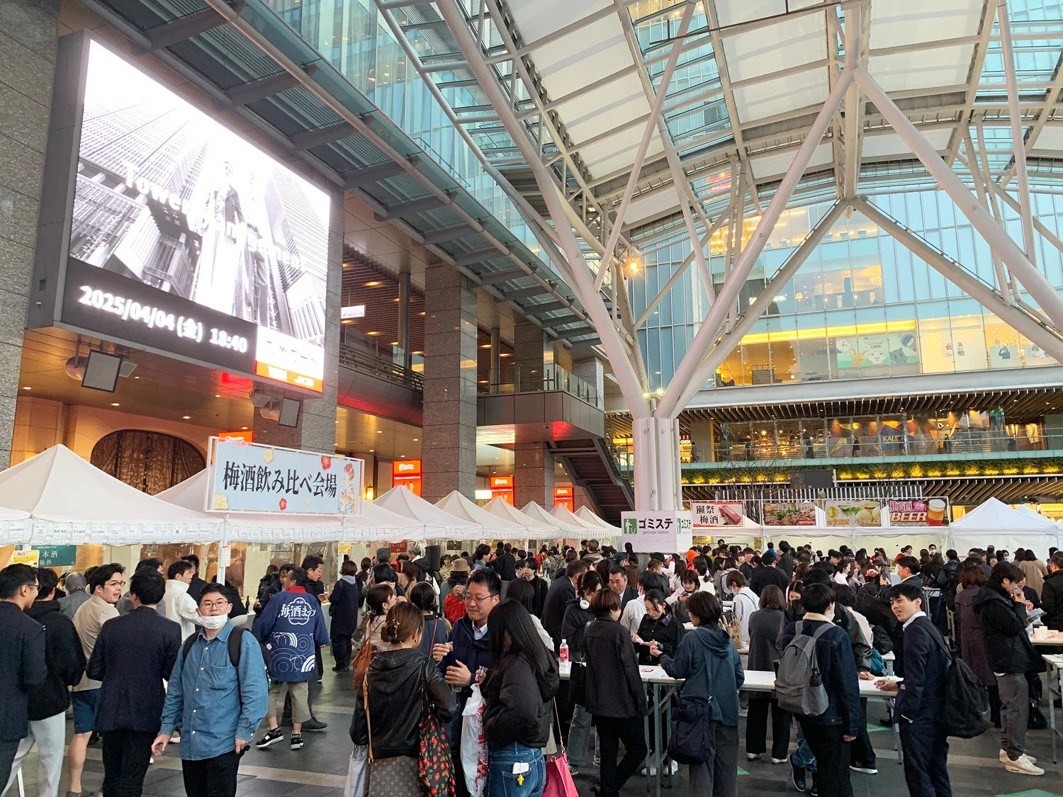 多くの人々で賑わう屋外イベント会場の様子。現代的な建築物の下で梅酒の飲み比べや飲食のブースが並び、活気ある雰囲気です。