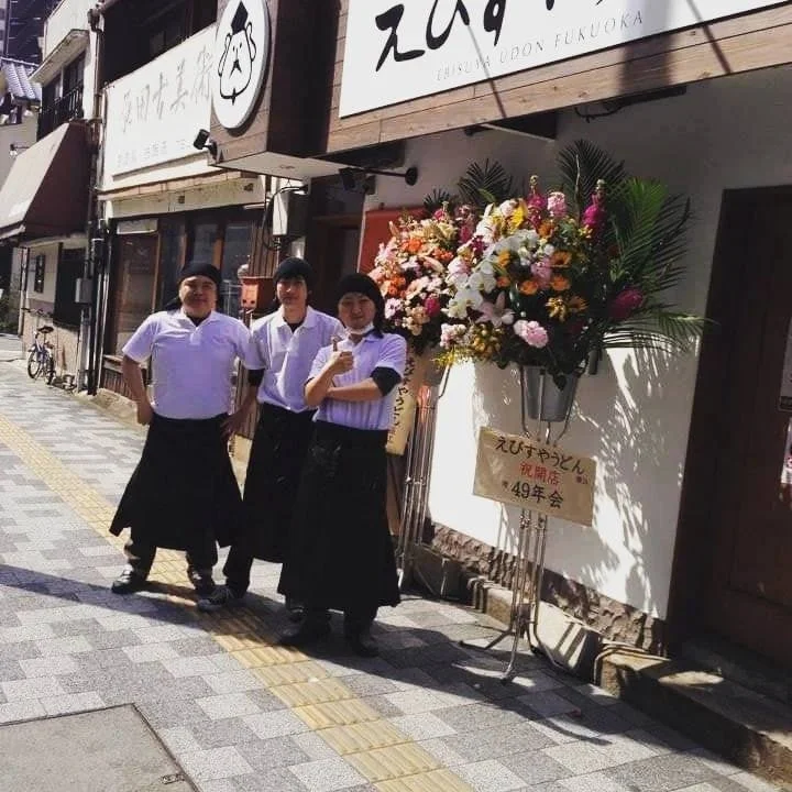 「えびすやうどん福岡」の店先で開店祝いの花と共に立つ3人の店員