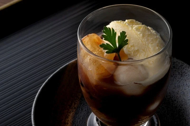 グラスに入ったコーヒーフロート。冷たいコーヒーの上にバニラアイスクリームが乗り、黄金色のトッピングと緑のハーブが添えられている