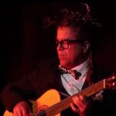 暗い背景の中、スーツと蝶ネクタイを着用し眼鏡をかけた男性がアコースティックギターを演奏しています。顔とギターに赤い光が当たっており、演奏中の真剣な表情が伺えます。