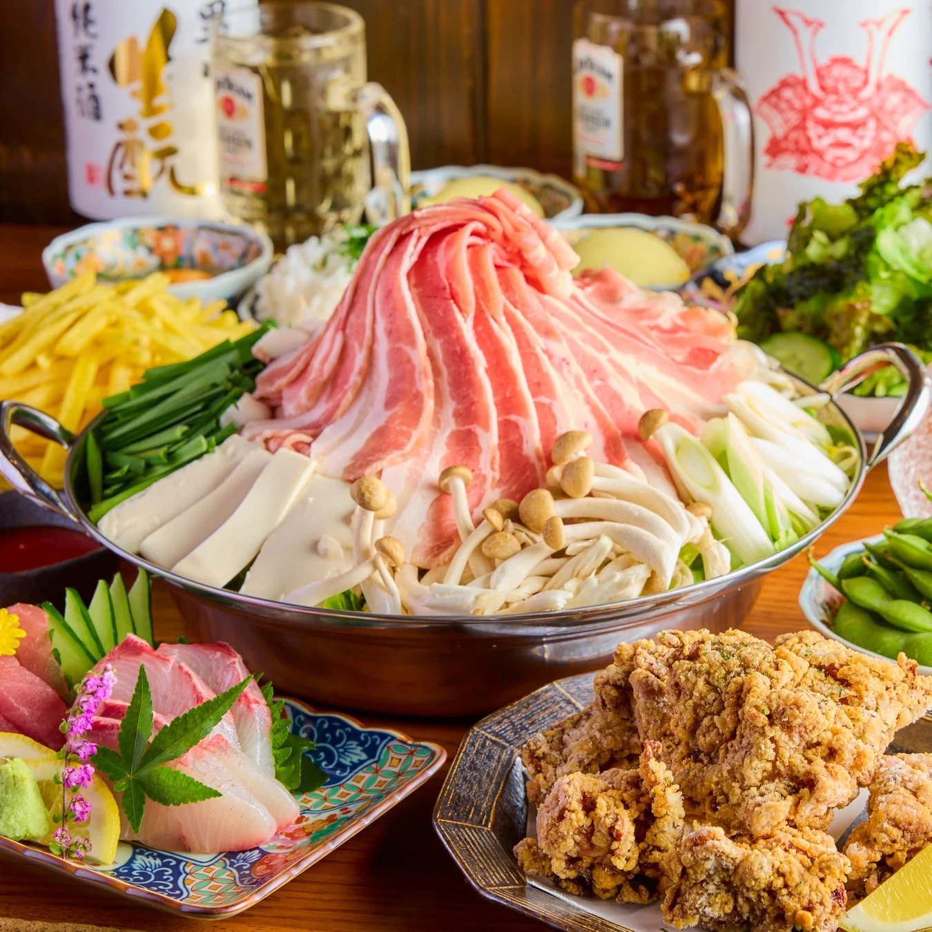 中央に豚肉と野菜がたっぷりの鍋が置かれ、周りには刺身、唐揚げ、枝豆などが並ぶ豪華な和食の食卓です