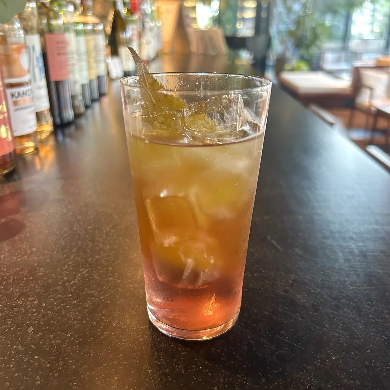 バーカウンターに置かれた、氷とミントの葉らしきガーニッシュが入ったカクテルのクローズアップ。背景には様々なボトルが並んでおり、おしゃれなバーの雰囲気を伝えています。