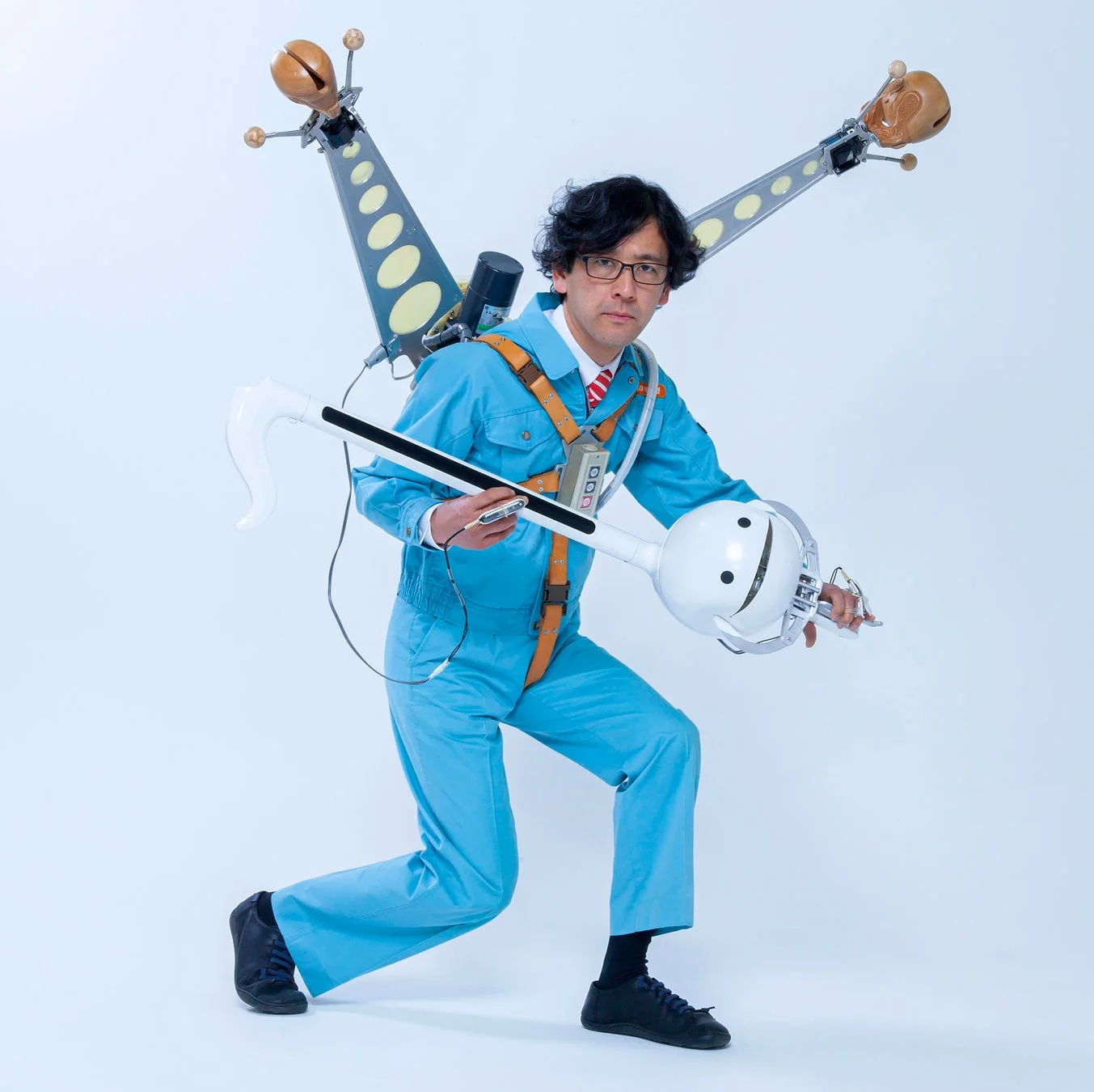 青い作業着を着た男性が、背中に奇妙な装置を背負い、巨大なオタマトーンのような楽器を抱えてポーズをとっている。全体的にユニークで創造的な印象を与える画像。