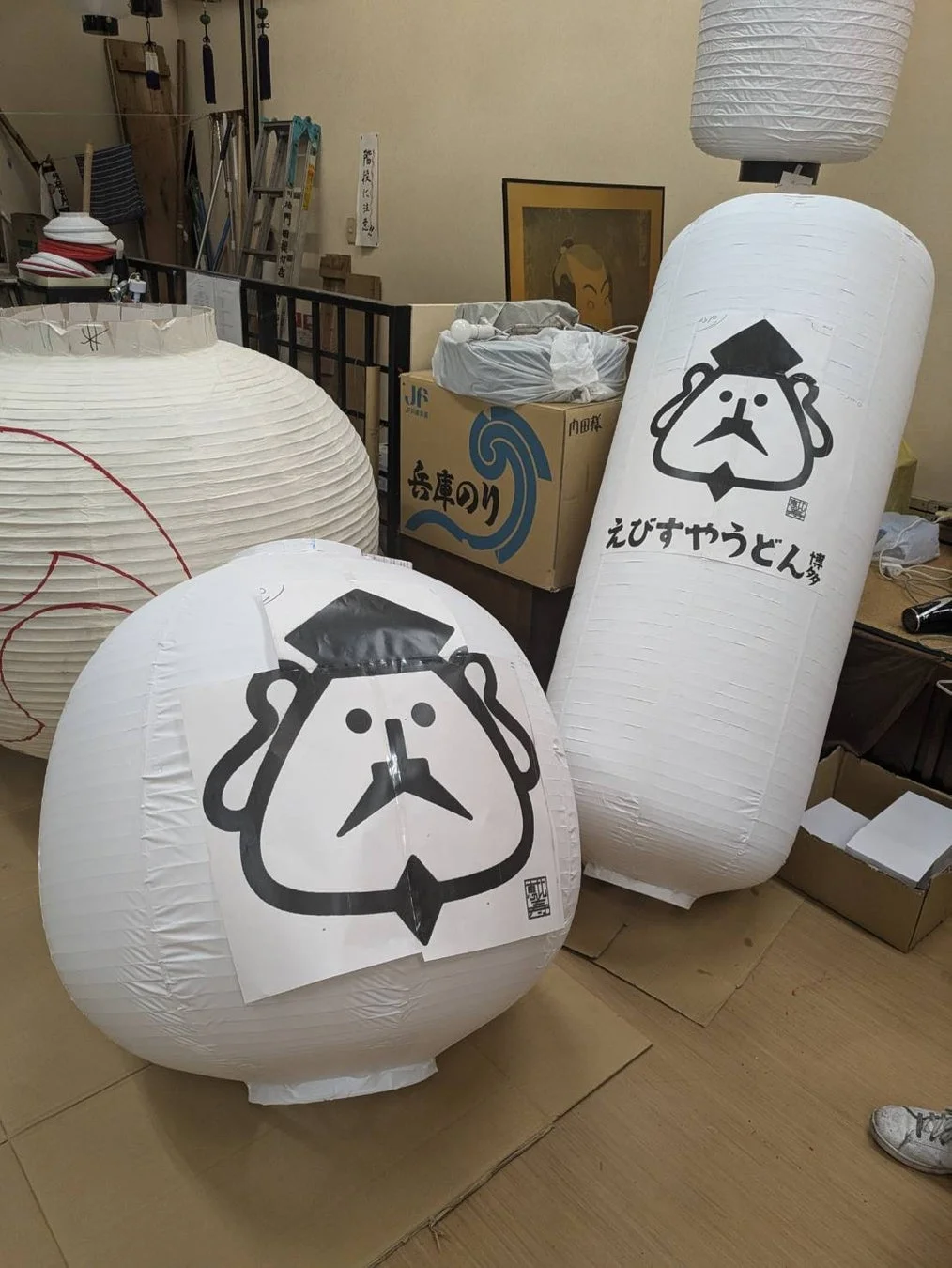 「えびすやうどん」と書かれた、えびす様のようなキャラクターが描かれた提灯が並ぶ作業場