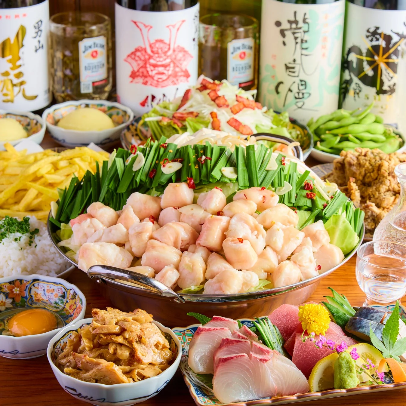 もつ鍋を中心に、刺身の盛り合わせ、フライドポテト、枝豆、唐揚げなど様々な居酒屋料理が並び、日本酒やジムビームのボトルも見える豪華な宴会の食卓風景