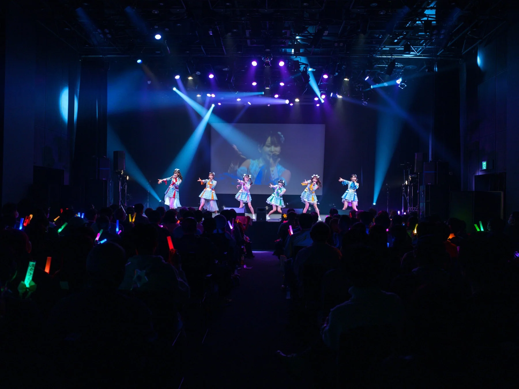 複数の女性アイドルグループがステージでパフォーマンスしており、大勢の観客がカラフルなサイリウムを振って応援しているライブコンサートの様子