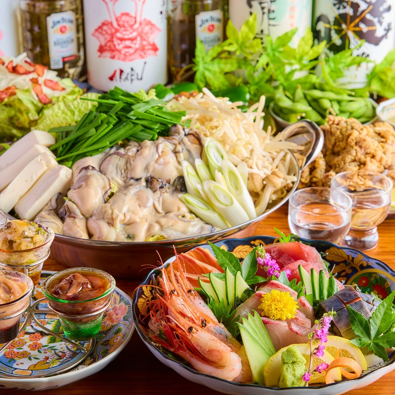 豪華な和食の宴会料理が並ぶ食卓。牡蠣や野菜たっぷりの鍋、新鮮な刺身盛り合わせ、唐揚げ、枝豆などが所狭しと置かれ、日本酒やウイスキーのボトルも見えます