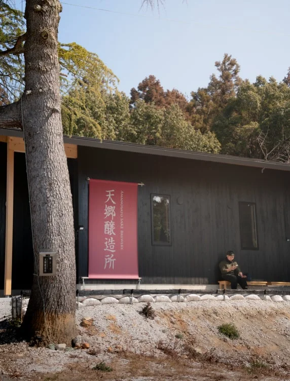 「天郷醸造所」の赤い看板がかかる黒い建物と、手前に立つ大きな木が特徴的な屋外の風景。建物前では一人の男性が椅子に座って休憩している。
