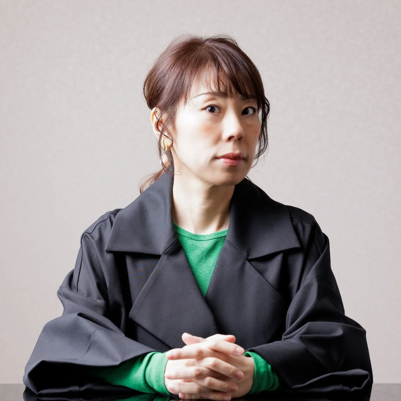 黒いアウターと緑色のインナーを着用した日本人女性が、室内で手を組んで座っているポートレートです。真剣な表情でカメラを見つめています。