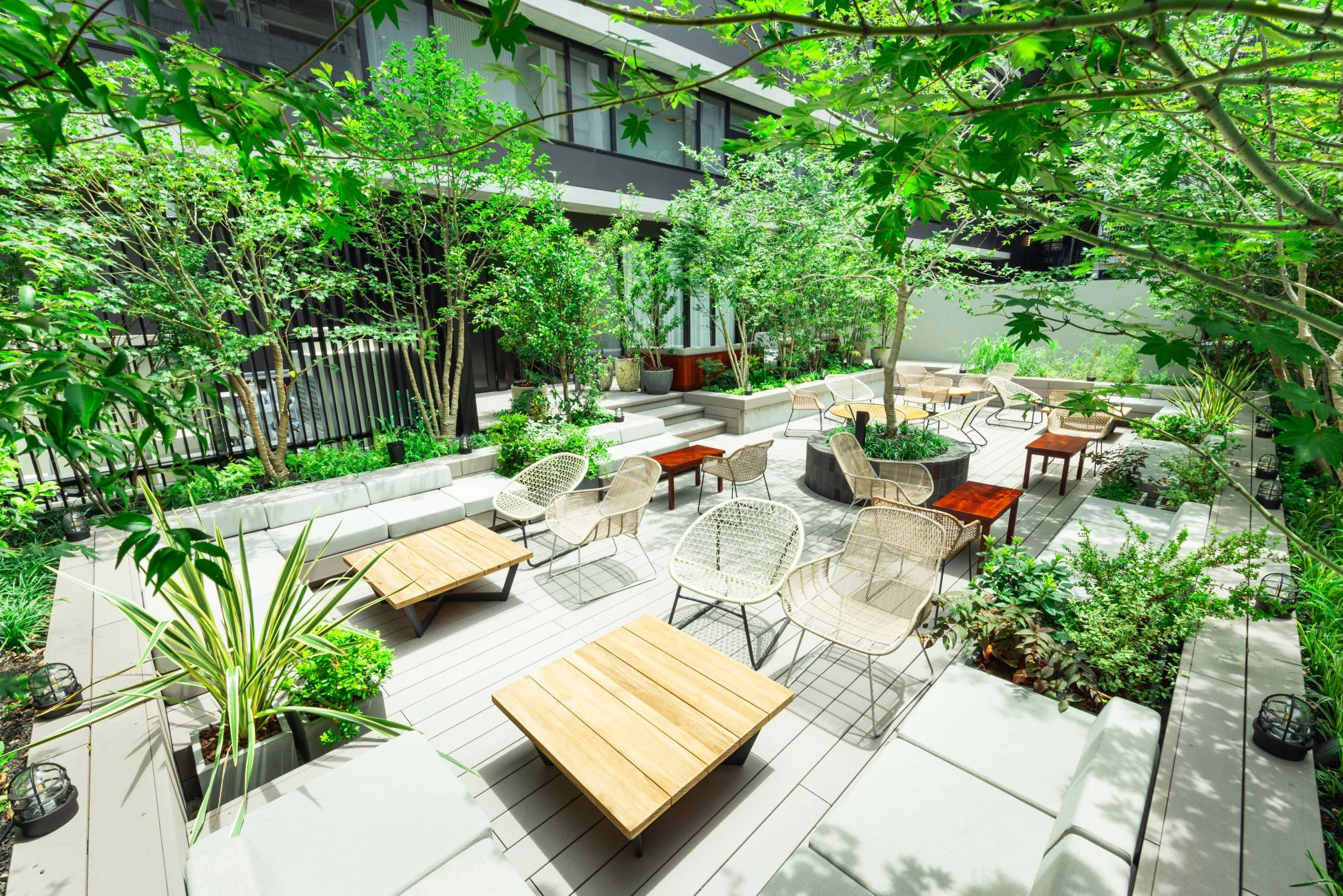 都会の中にある緑豊かな屋外テラス空間です。たくさんの木々や植物に囲まれ、モダンなデザインの椅子とテーブルが配置されており、リラックスできる雰囲気を提供しています。