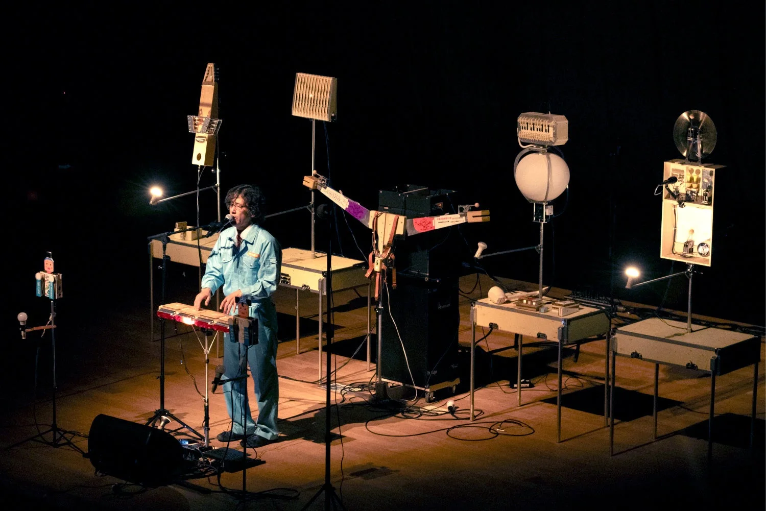 暗いステージで、青い作業着を着た男性が自作と見られるユニークな楽器の数々を使って演奏している様子。スポットライトが演者と複雑な機材を照らしています。