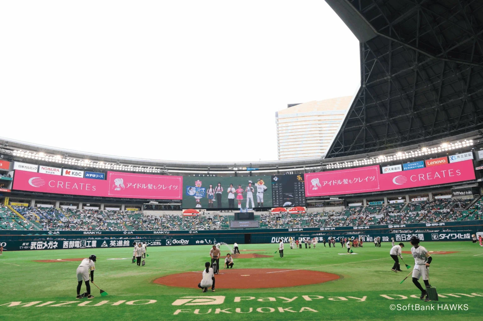 野球場でグラウンド整備が行われている様子、大型ビジョンには広告が映し出されている