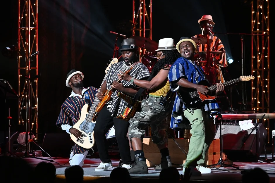 ステージ上で数人の男性バンドがギターやベースを演奏しながらライブパフォーマンスを行っています。彼らは特徴的なストライプ柄のシャツと帽子を身につけ、躍動感あふれる演奏を披露しています。