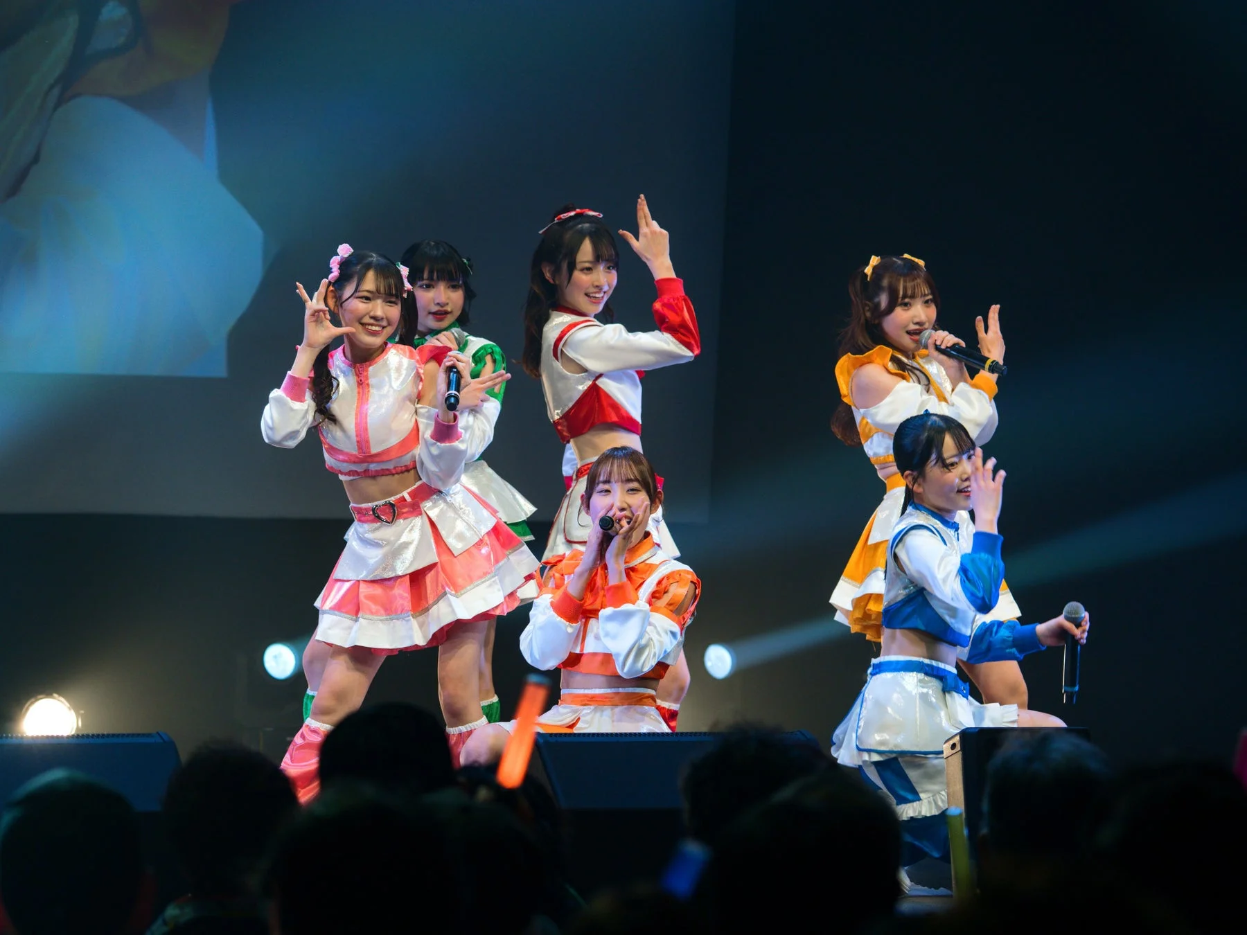 カラフルな衣装を身につけた複数の女性アイドルがステージ上で歌とダンスを披露しているライブパフォーマンスの様子