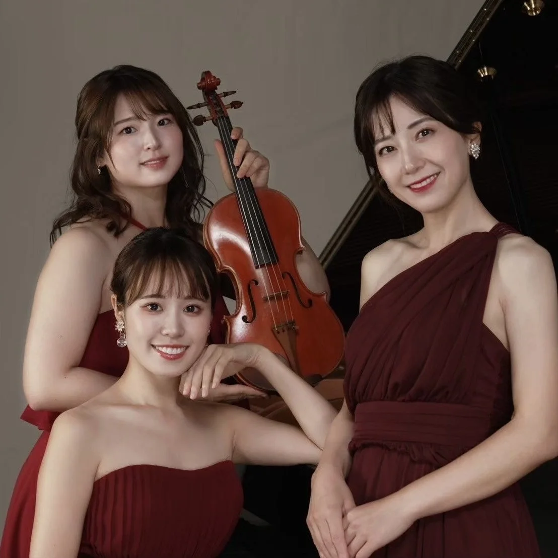 ワインレッドのドレスをまとった3人の女性が笑顔でポーズをとっています。中央の女性はバイオリンを持ち、背景にはピアノの一部が見え、クラシック音楽の演奏家グループのような雰囲気のポートレート写真です。