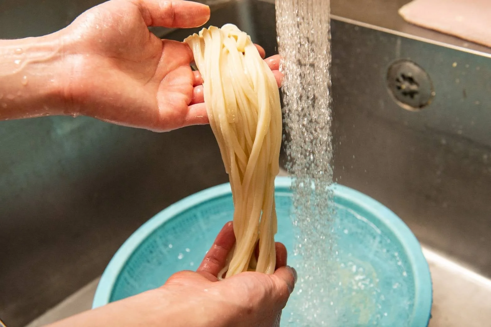 キッチンシンクで、手がうどんのような太麺を流水で洗っている場面