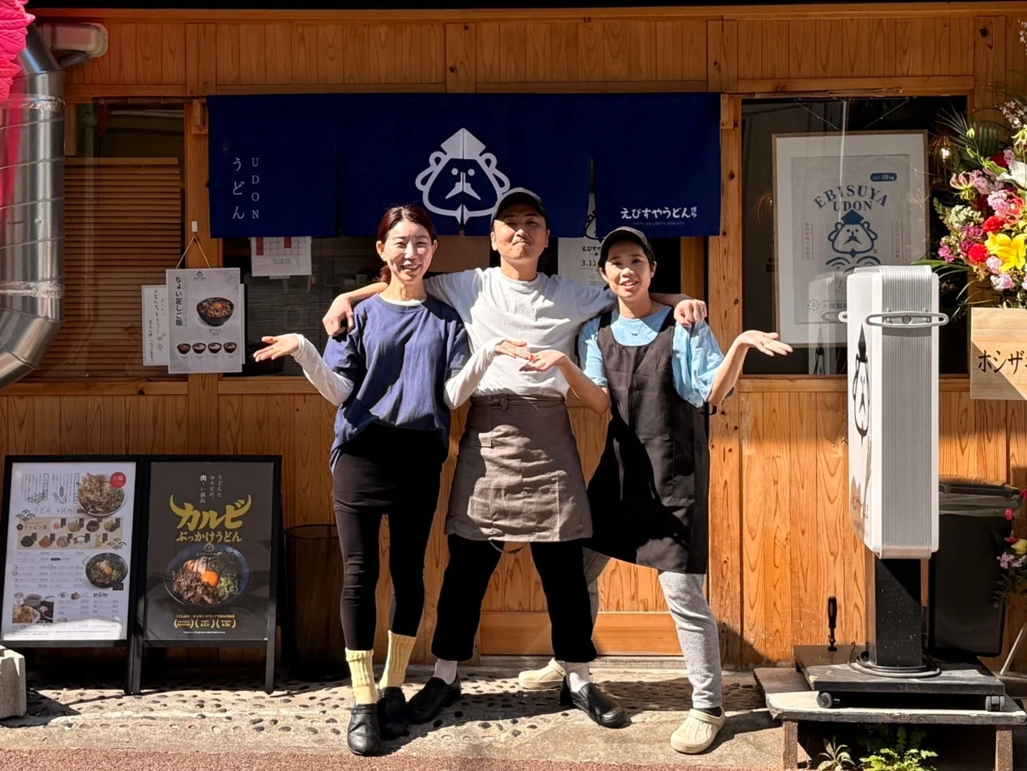 晴れた日に、うどん店の店先で笑顔の店員らしき3人がポーズをとっている