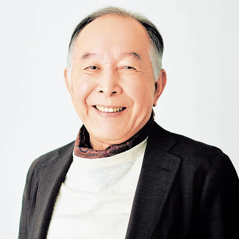 白背景の前で、親しみやすい笑顔を見せるアジア系のシニア男性のポートレートです。ダークなジャケットとライトなセーター、そして模様入りのスカーフを着用しています。