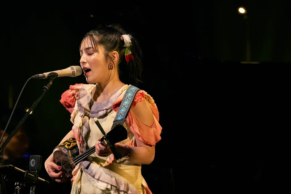 民族衣装をまとった女性歌手が、ステージ上でマイクに向かって歌いながら三線を演奏している様子