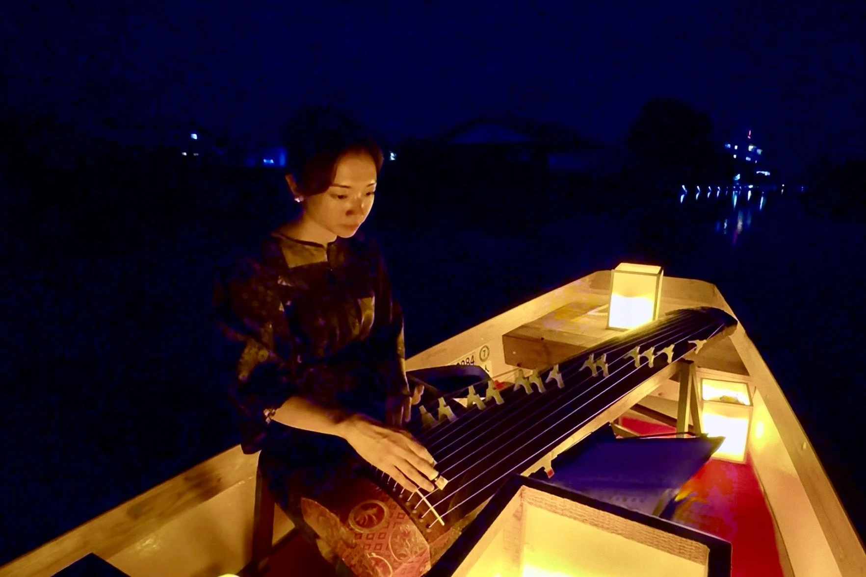 夜の川を屋形船で進む中、和服姿の女性が琴を演奏している
