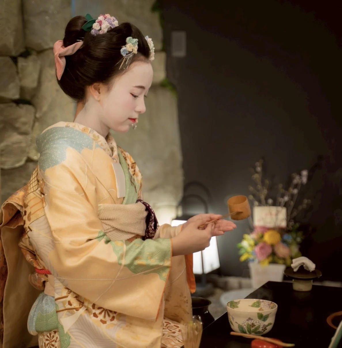 着物と白塗りの化粧をした舞妓が、茶碗と柄杓を用いて茶道を披露している様子。日本の伝統文化と優雅さを感じさせる瞬間を捉えた一枚です。