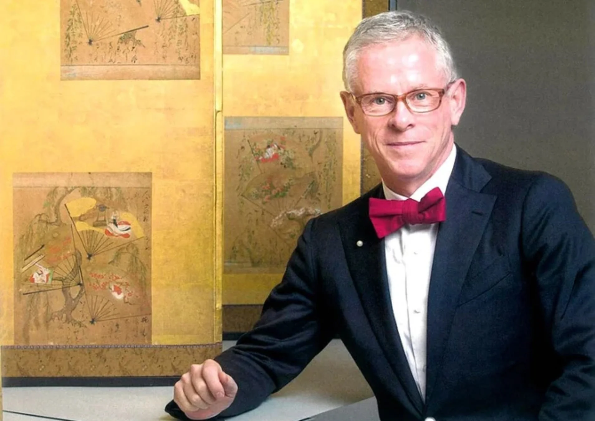 金屏風を背景に、ダークスーツに赤い蝶ネクタイと眼鏡をかけた白髪の男性