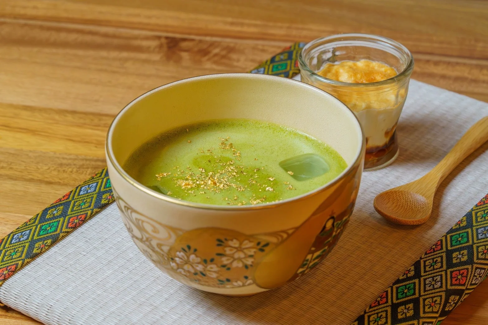 金箔が散りばめられた抹茶と、隣に置かれたデザート、木製のスプーンが写っています。和風のマットの上に置かれ、落ち着いた雰囲気の食卓を演出しています。