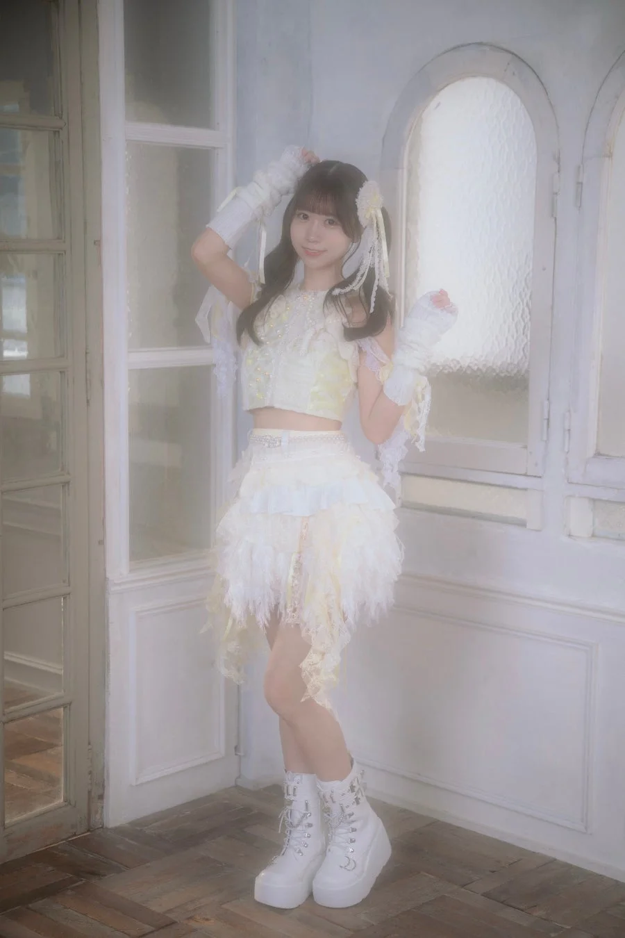 白いレースとフェザーの衣装を身につけた女性アイドルが、明るいスタジオで微笑んでいる全身ポートレート。ツインテールと厚底ブーツが特徴的で、夢のような雰囲気を醸し出しています。