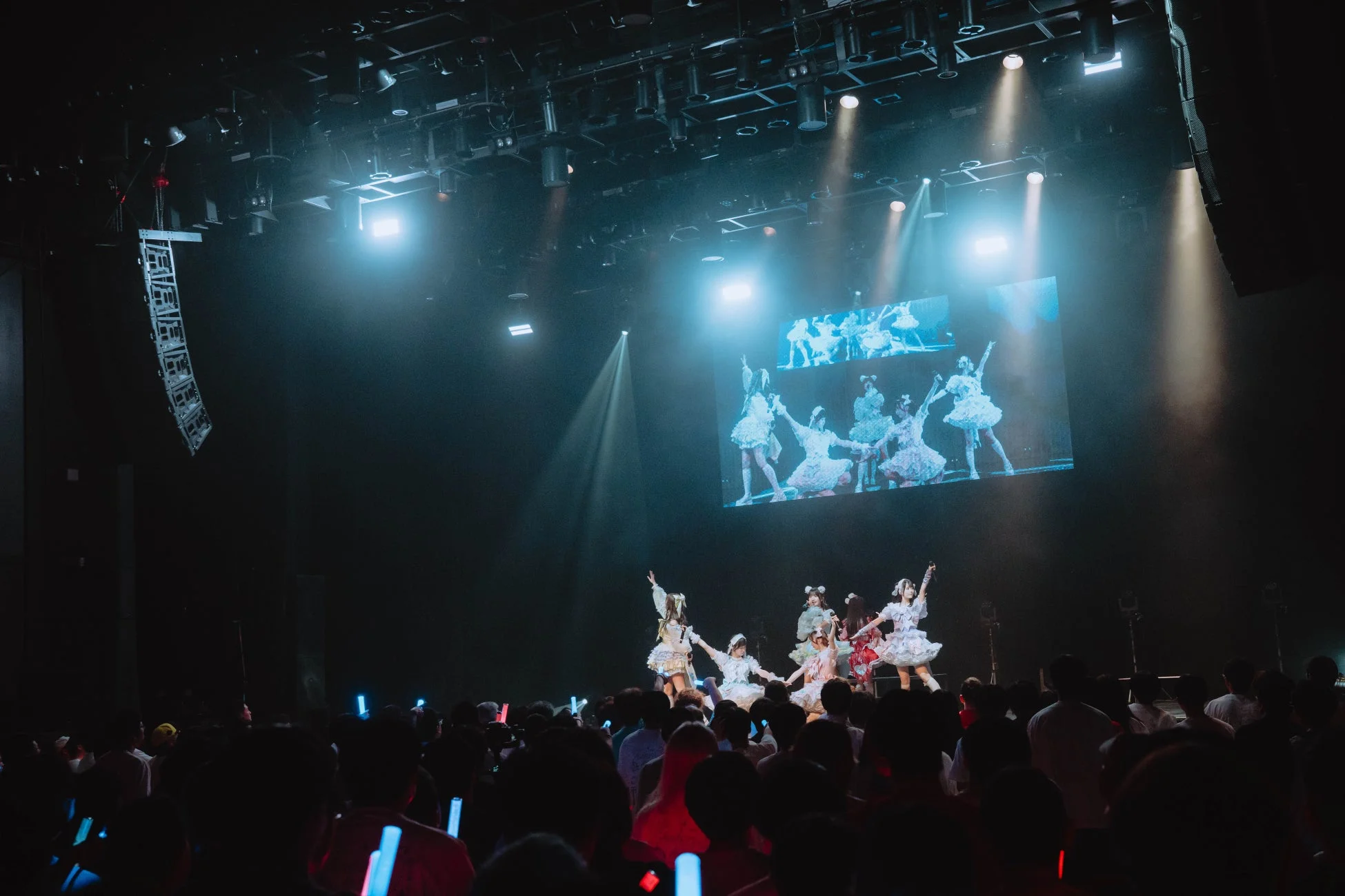 暗い会場で行われているライブコンサートの様子です。ステージ上では数人のパフォーマーが踊っており、後方の大型スクリーンにも映像が映し出されています。観客はペンライトを振って盛り上がっています。