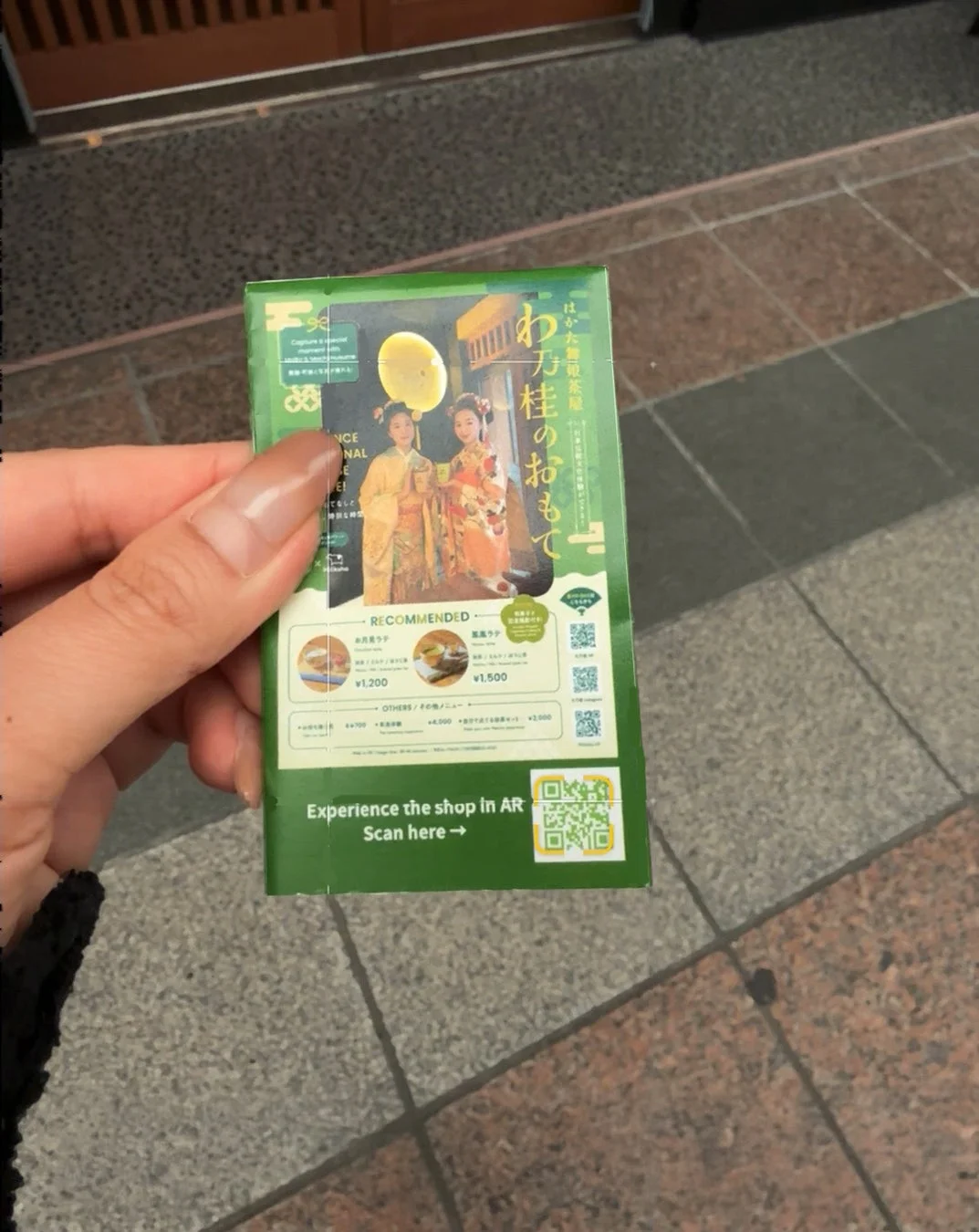 はかた伝統茶屋の緑色のパンフレットを手に持っている画像。和装の女性二人、お月見ラテや黒糖ラテなどのメニュー、AR体験へのQRコードが掲載されている。