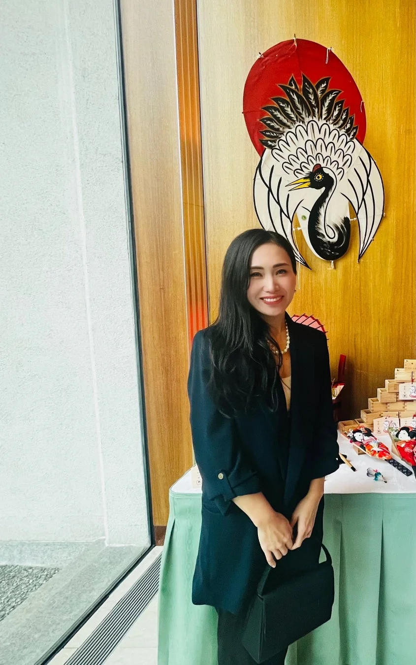 長い黒髪の女性が笑顔で立っており、背景には鶴と日の丸をモチーフにした和風の装飾が見えます