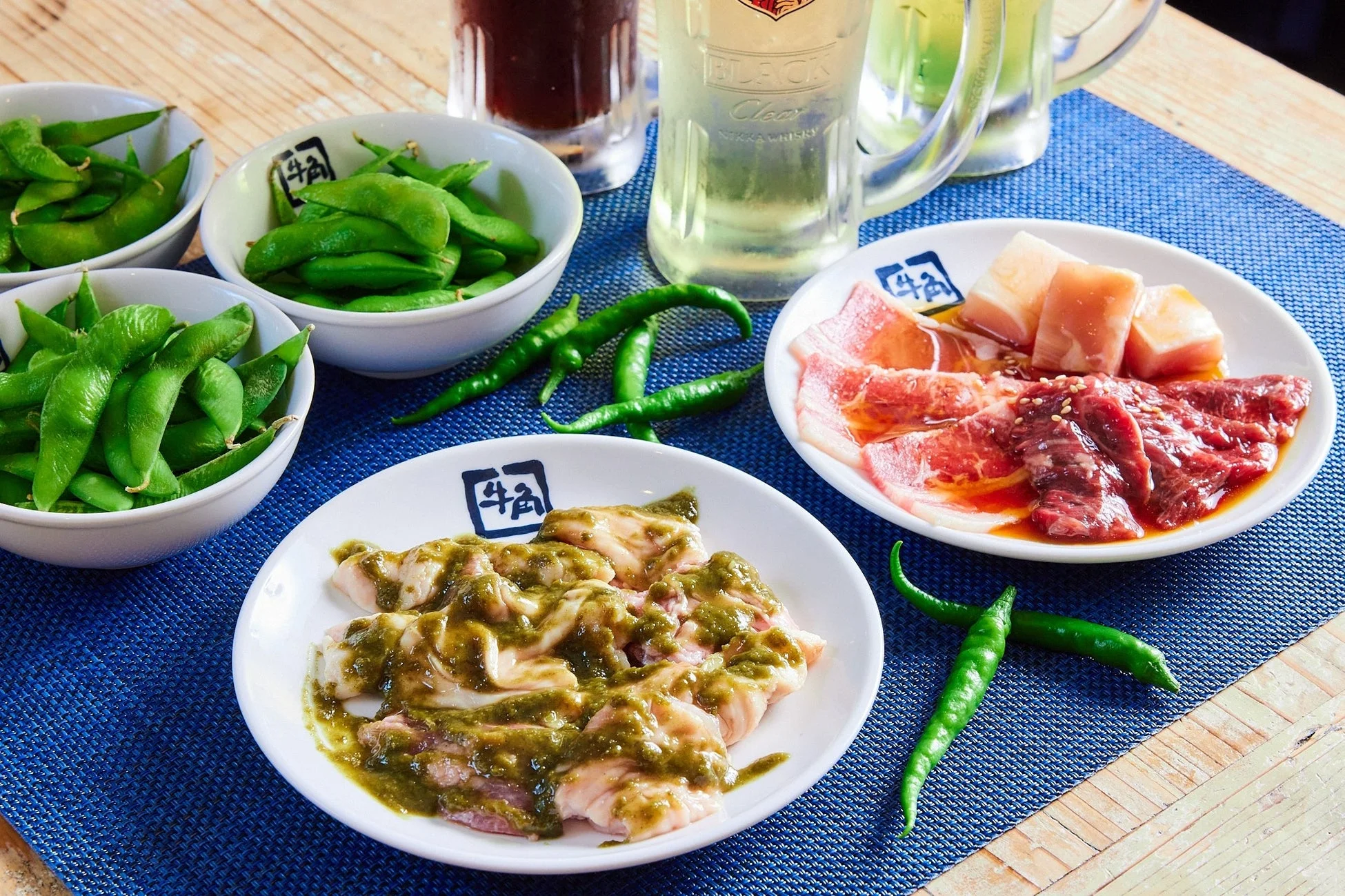 焼肉の準備がされた食卓の風景。牛角のロゴ入り皿に盛られた生肉や、緑色のソースがかかった肉、枝豆が並びます。ビールやハイボールのグラス、緑の唐辛子も添えられ、賑やかな食事のひとときを伝えています。