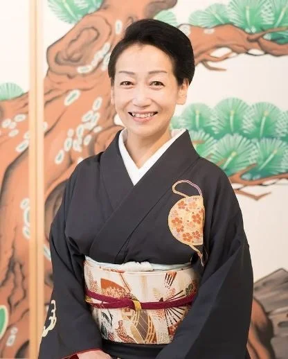 笑顔の日本人女性が、黒地の着物に華やかな帯を締めて立っている。背景には松の木が描かれた和風の絵画があり、伝統的な雰囲気を醸し出している。