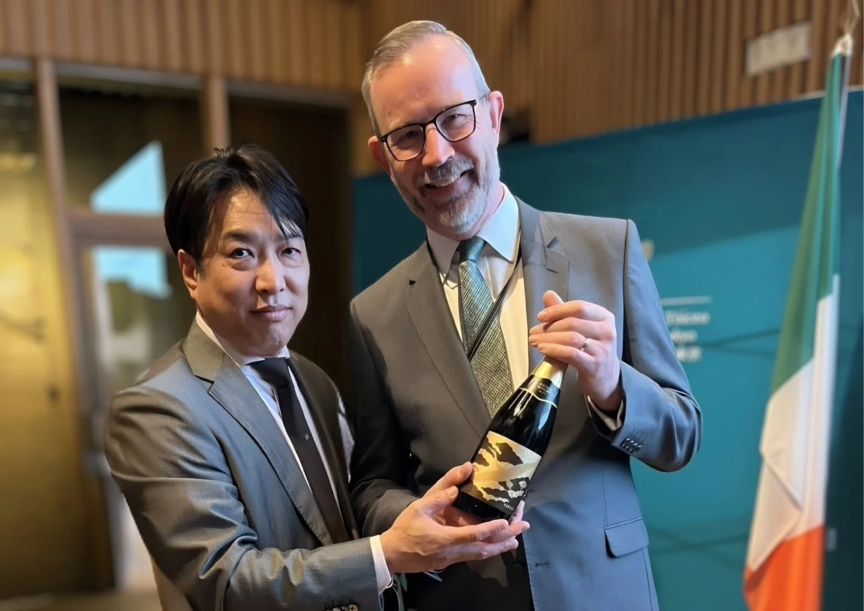 スーツ姿の2人の男性が笑顔でカメラに向かい、1本のボトルを共有して持っています