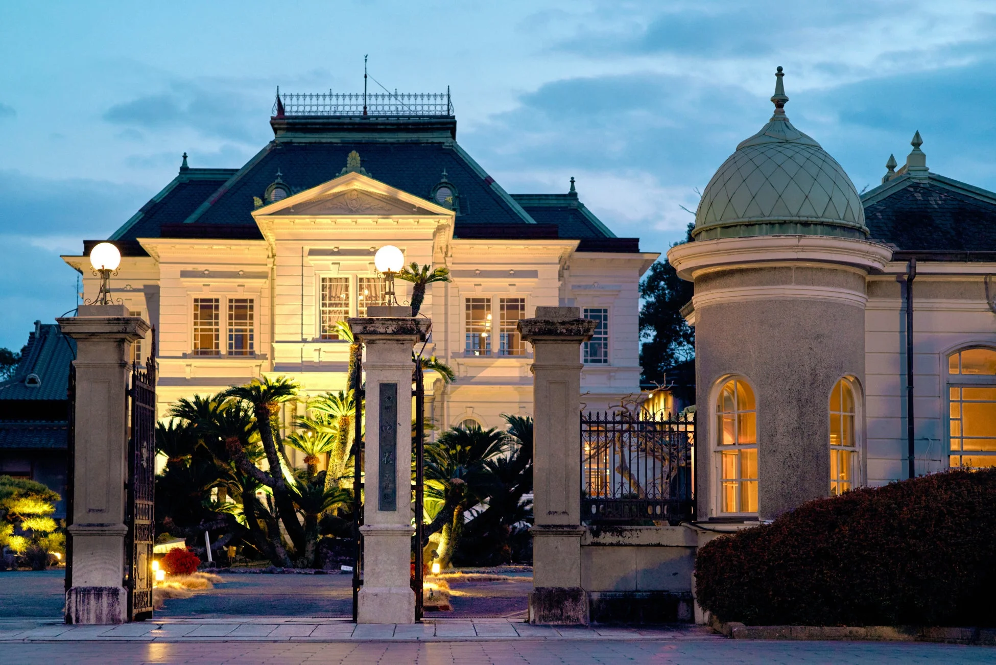 夕暮れ時にライトアップされた、壮麗な洋館と庭園の様子
