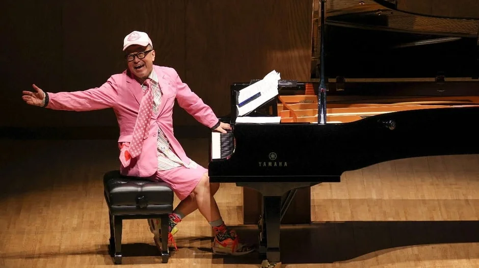 ピンクのスーツとキャップを身につけた男性が、木製のステージで黒いグランドピアノに向かい、腕を広げて満面の笑みを浮かべている