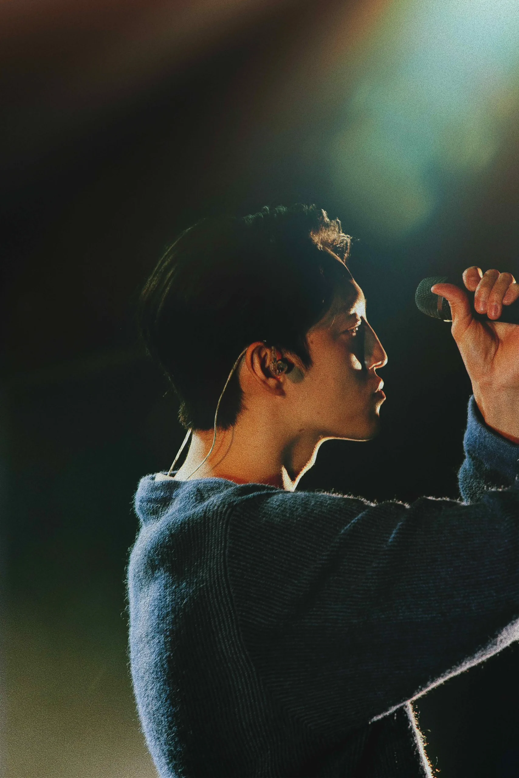 暗いステージでスポットライトを浴びながらマイクを握り歌う男性の横顔。真剣な表情でパフォーマンスに集中している様子がうかがえる。